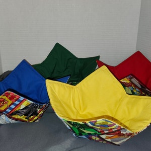 May include: Four fabric bowl cozies in blue, green, red, and yellow. Each bowl has a solid color interior and a patterned exterior with comic book-style graphics. The cozies are designed to hold bowls of hot food.