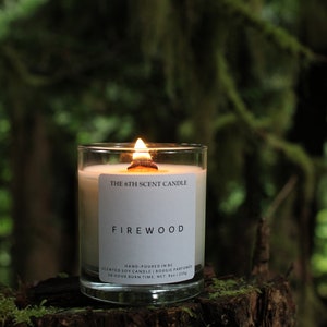 May include: A white soy candle in a clear glass jar with a wooden wick. The candle is labelled "The 6th Scent Candle" and "Firewood". The label also states "Hand-poured in BC", "Scented Soy Candle | Bougie Parfumée", and "50 Hour Burn Time. Net. 9oz | 255g".