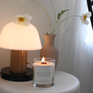 May include: A clear glass candle jar with a white label reading THE 6TH SCENT CANDLE EUCALYPTUS + SPEARMINT, featuring a lit wooden wick. The candle sits beside a mushroom-shaped lamp with a white glass shade and a tan base, and a beige ceramic vase.