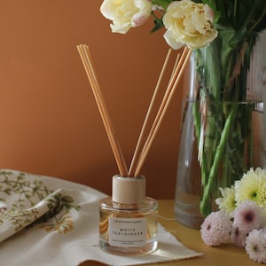 May include: A clear glass diffuser bottle with a wooden cap and reeds. The bottle is filled with a light yellow liquid and has a label that reads "The Sixth Scent Candle White Tea + Ginger Hand-Poured Reed Diffuser 100ml e." The bottle is sitting on a white and yellow floral tablecloth.