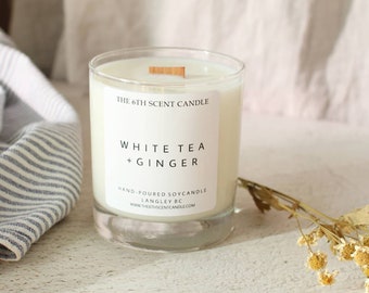 May include: A white soy candle in a clear glass jar with a wooden wick. The label reads "THE 6TH SCENT CANDLE", "WHITE TEA + GINGER", "HAND-POURED SOYCANDLE", "LANGLEY BC", and "WWW.THESTHSCENTCANDLE.COM".