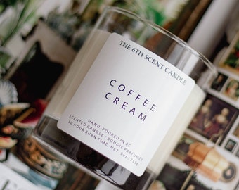 Op de afbeelding: Een glazen kaars pot met een wit label waarop staat "THE 6TH SCENT CANDLE COFFEE CREAM HAND-POURED IN BC SCENTED CANDLE | BOUGIE PARFUMÉE 50 HOUR BURN TIME. NET. 9oz | 255g".
