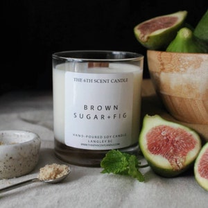 May include: A clear glass candle jar with a white label that reads "THE 6TH SCENT CANDLE", "BROWN SUGAR+FIG", "HAND-POURED SOY CANDLE", "LANGLEY BC", and "WWW.THE6THSCENTCANDLE.COM". The candle is surrounded by figs and a sprig of mint.