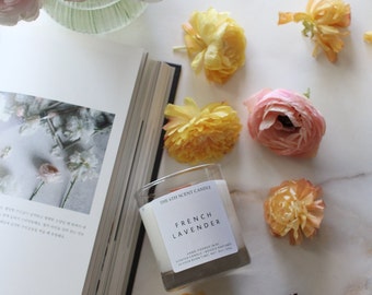 Op de afbeelding: Een witte kaars pot met een zwart label waarop staat "The 6th Scent Candle French Lavender" en "Hand-poured wax, Scented candle, 8 oz, Soy wax, 18 hour burn time, Net wt 227g". De kaars is omringd door gele en roze bloemen op een wit marmeren oppervlak.