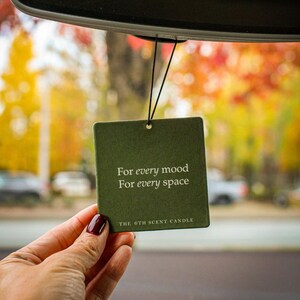 Puede incluir: Un ambientador de coche verde con el texto "For every mood For every space" y "THE 6TH SCENT CANDLE" colgado del espejo retrovisor de un coche.