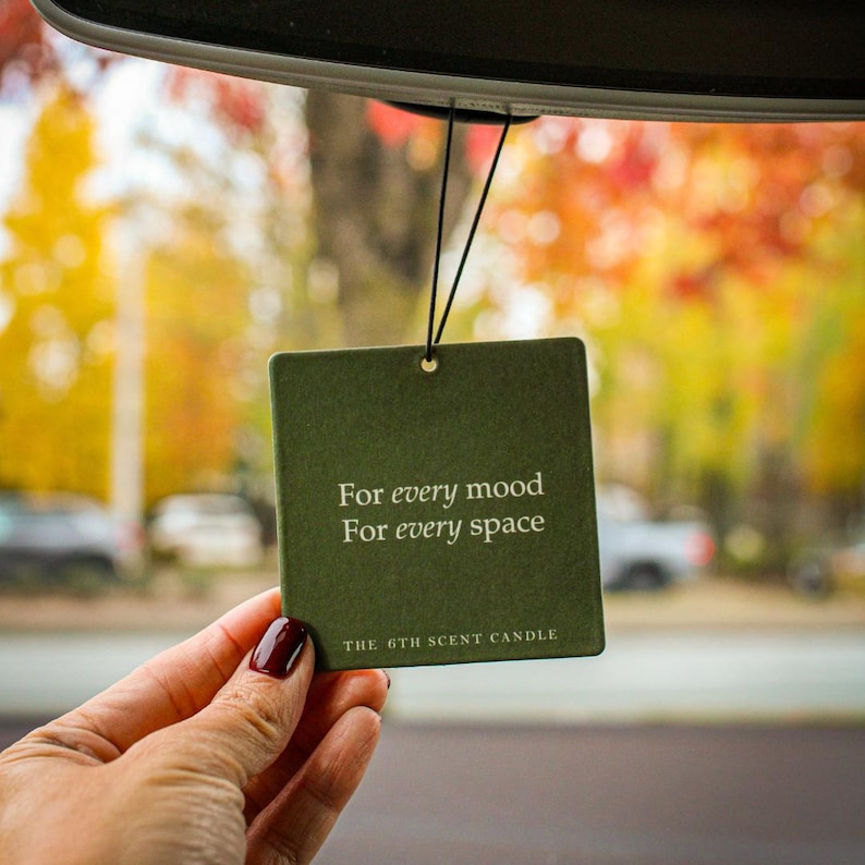 Puede incluir: Un ambientador de coche verde con el texto "For every mood For every space" y "THE 6TH SCENT CANDLE" colgado del espejo retrovisor de un coche.