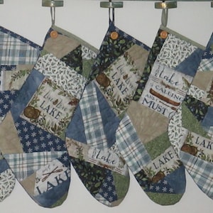 May include: Five patchwork Christmas stockings featuring blue, green, and tan fabric panels with lake-themed prints. Each stocking includes a hanging loop with a wooden button and displays text such as The Lake Is My Happy Place and Lake Love.