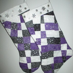 May include: Two quilted Christmas stockings with a patchwork design in purple, black, white, and silver. The top cuff is white with silver snowflake accents. The stockings are hanging against a white background.