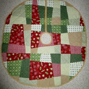 May include: A round, quilted Christmas tree skirt with a patchwork design. The skirt features squares of red, green, and tan fabrics with holiday patterns. A circular opening is in the center, and the edge is trimmed with a gold-colored binding.