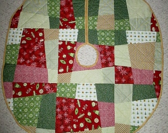 May include: A round, quilted Christmas tree skirt with a patchwork design. The skirt features squares of red, green, and tan fabrics with holiday patterns. A circular opening is in the center, and the edge is trimmed with a gold-colored binding.