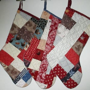 May include: Three quilted Christmas stockings with patchwork designs. Each stocking features a variety of fabrics in red, white, blue, and brown, with patterns including stars, chickens, and paisley. They hang from small loops.