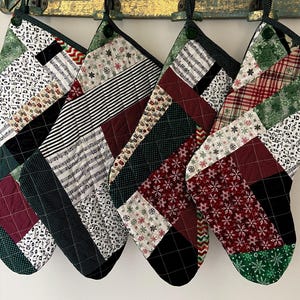 May include: Four handmade Christmas stockings with various patchwork patterns. Designs include musical notes, stripes, snowflakes, and plaid. Each stocking has a green loop for hanging and a black border.