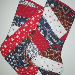 May include: Two quilted Christmas stockings with red, white, and blue patchwork. One stocking features red fabric with white stars, while the other has a football pattern. Both have red hanging loops and the word "Patriots".