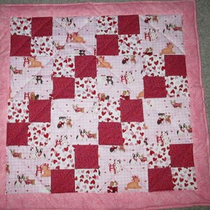 May include: A handmade patchwork quilt with a pink border. The quilt features a pattern of alternating red squares and squares with a print of dogs and hearts on a pink background. The quilt is likely made of cotton and is suitable for a baby or pet.