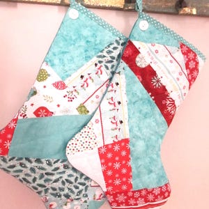 May include: Two handmade Christmas stockings. Each stocking features a patchwork design with teal, red, and white fabrics. The patterns include snowflakes, snowmen, and festive greenery. Each stocking has a teal hanging loop and a white button.
