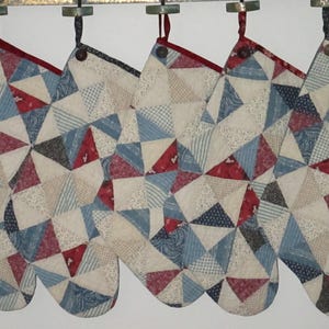May include: Five quilted Christmas stockings hanging in a row, each featuring a patchwork design of triangles in shades of red, white, and blue. The stockings are constructed from various patterned fabrics and are suspended from a horizontal wooden rack.