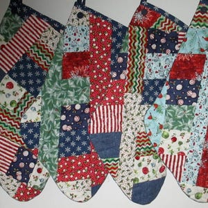 May include: Four Christmas stockings with patchwork designs. Each stocking features a variety of fabrics in red, white, blue, and green, with patterns including stripes, snowflakes, and holiday motifs. The stockings have denim cuffs and hanging loops.