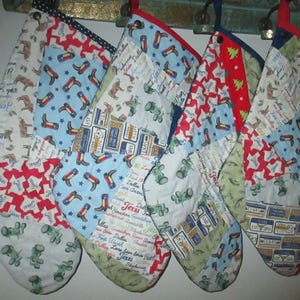 May include: Five quilted Christmas stockings with various patterns, including cowboy boots, tractors, and Texas-themed designs. Each stocking has a different color scheme, with red, blue, and green accents. They are hanging from a decorative metal bar.