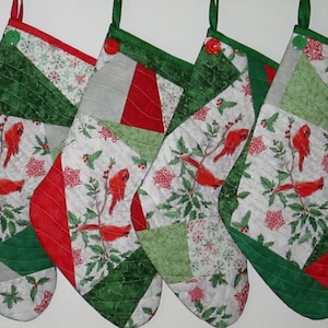 May include: Four quilted Christmas stockings featuring a patchwork design of red cardinals, green holly leaves, and white snowflake patterns. Each stocking is accented with a solid red or green fabric border and a single button at the top hanging loop.
