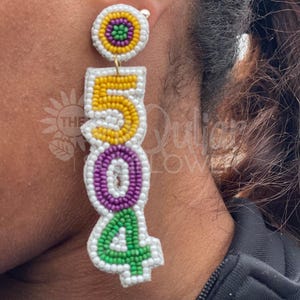 Puede incluir: Un par de pendientes de cuentas con una base blanca y cuentas de colores. Los pendientes tienen forma de los números 504. Las cuentas son amarillas, moradas y verdes.