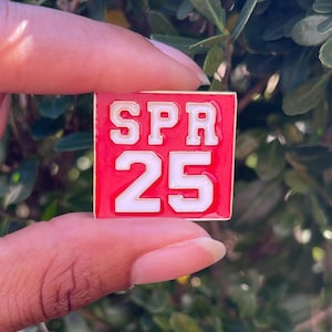 May include: A red enamel pin with a gold border. The pin features white text that reads "SPR 25".