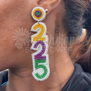 Puede incluir: Un par de pendientes de cuentas con fondo blanco y cuentas de colores en forma de los números 2, 2, 5. Las cuentas son amarillas, moradas y verdes.