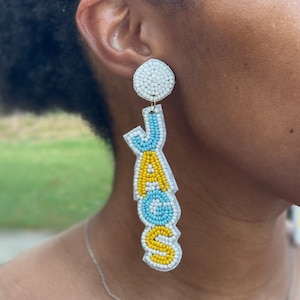 May include: A pair of beaded earrings spelling out the word "YAS" in blue, yellow, and white beads. The earrings are hanging from a person's ear.