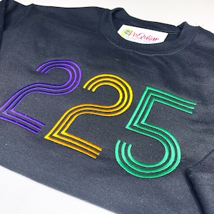 Puede incluir: Sudadera negra con el número "225" bordado en morado, dorado y verde. La sudadera tiene cuello redondo y una etiqueta que dice "PeQuliar Sunflower".