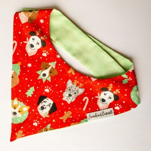 May include: A red dog bandana with a festive Christmas pattern. The design features various dog breeds, Christmas trees, paw prints, and gingerbread men. The bandana has a light green lining and a small label that reads "Frankie & Emmett."