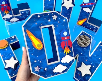 May include: A set of blue, white, and red cardboard letters spelling out the word "COOL" with a space-themed design. The letters are decorated with stars, rockets, comets, and clouds. The letters are standing on a blue surface.
