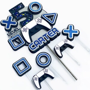 May include: A set of blue and silver cake toppers featuring a video game controller, a square, a circle, a triangle, an X, a plus sign, the number 5, and the text "Level" and "Carter".