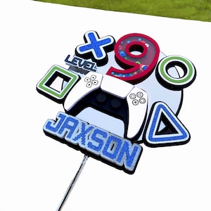 May include: A blue and silver cake topper with the name Jaxson, a video game controller, a blue triangle, a green square, a red number 9 with glitter, a blue X, and the words "Level" on it.