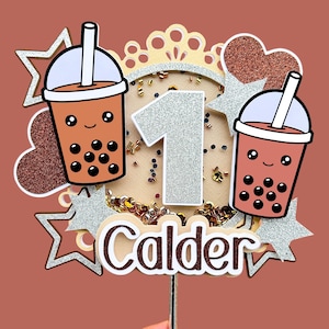 May include: A cake topper featuring a silver glitter number one, two brown and white bubble tea cups with faces, a brown heart, two silver glitter stars, and the name "Calder" in brown letters.