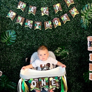 Pode incluir: Uma crian&ccedil;a senta-se numa cadeira alta decorada com uma faixa que diz "ONE". Uma faixa acima diz "HAPPY BIRTHDAY". O fundo &eacute; uma parede verde com folhas tropicais. A crian&ccedil;a usa uma camiseta branca com um gr&aacute;fico de dinossauro.