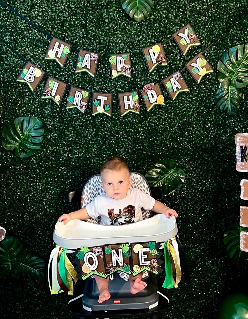 Pode incluir: Uma crian&ccedil;a senta-se numa cadeira alta decorada com uma faixa que diz "ONE". Uma faixa acima diz "HAPPY BIRTHDAY". O fundo &eacute; uma parede verde com folhas tropicais. A crian&ccedil;a usa uma camiseta branca com um gr&aacute;fico de dinossauro.