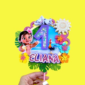 Puede incluir: Un adorno para pastel morado y rosa con un n&uacute;mero uno de purpurina, un personaje de dibujos animados de Moana, un gallo y el nombre "Eliana".