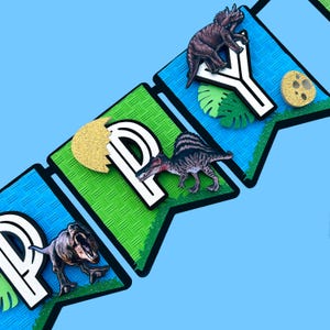 Pode incluir: Uma faixa com a palavra "HAPPY" com decora&ccedil;&otilde;es de dinossauros e folhas. As letras s&atilde;o brancas com contorno preto, em bandeirolas verdes e azuis. Detalhes em glitter dourado e fundo azul claro.