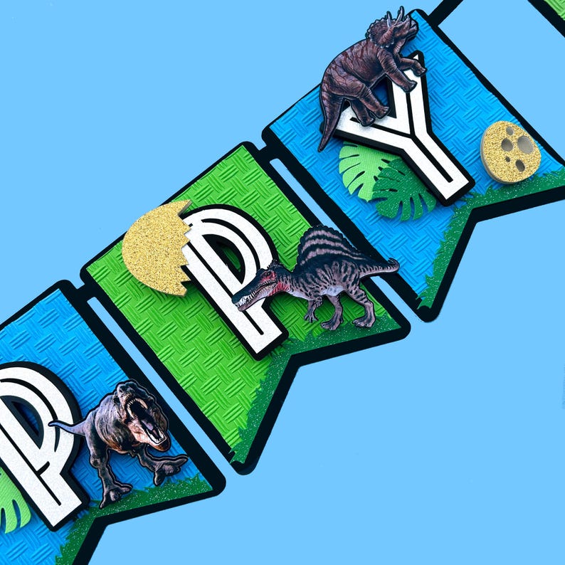 Pode incluir: Uma faixa com a palavra "HAPPY" com decora&ccedil;&otilde;es de dinossauros e folhas. As letras s&atilde;o brancas com contorno preto, em bandeirolas verdes e azuis. Detalhes em glitter dourado e fundo azul claro.