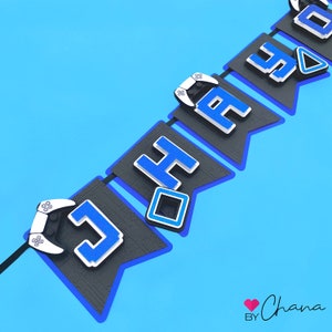 May include: A black and blue banner with the word "Happy" spelled out in blue letters. Each letter is on a separate flag with a white video game controller on the end of each flag. The banner is against a light blue background.