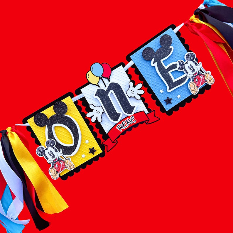 May include: A colorful banner with Mickey Mouse characters and the word "One" in black letters. The banner also includes the name "Rene" in red letters. The banner is decorated with yellow, red, blue, and white stars.