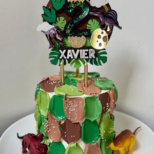 Pode incluir: Um bolo de anivers&aacute;rio decorado com cobertura verde e castanha, coberto com um topo de bolo com tema de dinossauro. O topo apresenta o n&uacute;mero "2", ilustra&ccedil;&otilde;es de dinossauros e o nome "XAVIER". Duas velas douradas s&atilde;o inseridas no bolo.