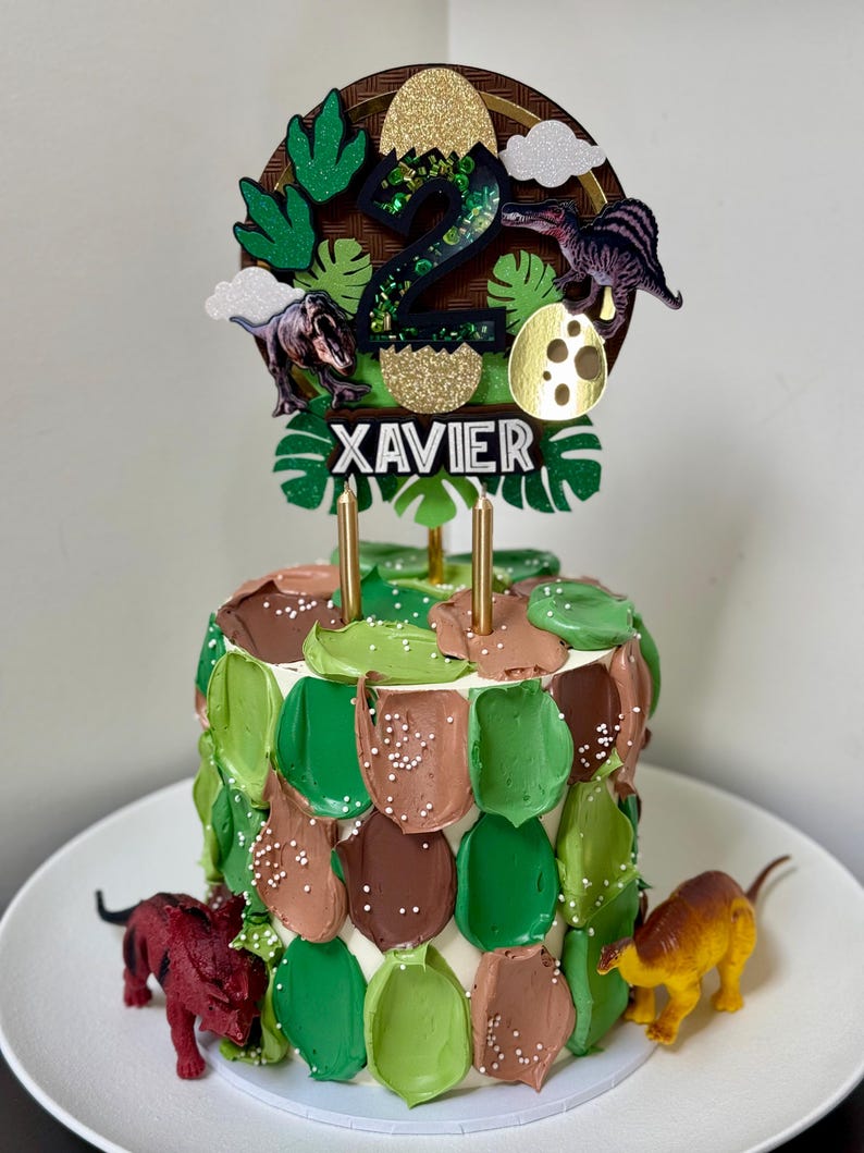 Pode incluir: Um bolo de anivers&aacute;rio decorado com cobertura verde e castanha, coberto com um topo de bolo com tema de dinossauro. O topo apresenta o n&uacute;mero "2", ilustra&ccedil;&otilde;es de dinossauros e o nome "XAVIER". Duas velas douradas s&atilde;o inseridas no bolo.