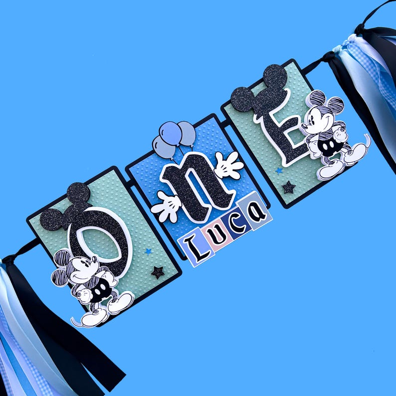 May include: A blue and green banner with the word "one" in black letters and a Mickey Mouse character on each end. The banner also includes the name "Luca" in colorful blocks.