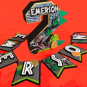 Pode incluir: Uma faixa de anivers&aacute;rio com tema de dinossauro com o n&uacute;mero 2 e o nome "Emerson". O n&uacute;mero &eacute; preto com uma textura de pedra e detalhes de glitter dourado. As pe&ccedil;as da faixa soletram "EMERSON".