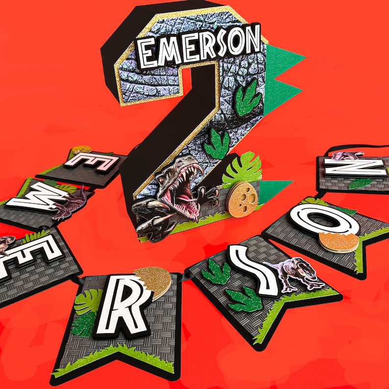 Pode incluir: Uma faixa de anivers&aacute;rio com tema de dinossauro com o n&uacute;mero 2 e o nome "Emerson". O n&uacute;mero &eacute; preto com uma textura de pedra e detalhes de glitter dourado. As pe&ccedil;as da faixa soletram "EMERSON".