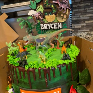 Pode incluir: Um bolo de anivers&aacute;rio decorado com o tema de dinossauros. O bolo verde tem gotas marrons e &eacute; coberto com dinossauros de brinquedo, glac&ecirc; verde e um topper com o nome "BRYCEN" e o n&uacute;mero "5".