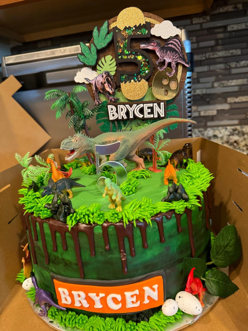 Pode incluir: Um bolo de anivers&aacute;rio decorado com o tema de dinossauros. O bolo verde tem gotas marrons e &eacute; coberto com dinossauros de brinquedo, glac&ecirc; verde e um topper com o nome "BRYCEN" e o n&uacute;mero "5".