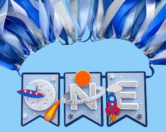 May include: A blue banner with the word "ONE" in silver glitter letters, decorated with space-themed elements. Includes a moon, a rocket, a sun, and a UFO. The banner is adorned with blue and silver ribbons, perfect for a first birthday celebration.