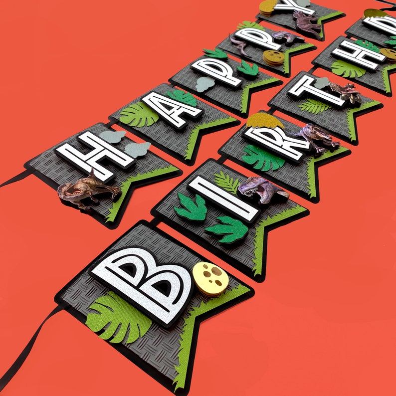 Pode incluir: Uma faixa com tema de dinossauro com a frase "HAPPY BIRTHDAY" e fita preta. Cada bandeira apresenta um padr&atilde;o de diamante cinza, detalhes verdes, figuras de dinossauros e recortes de folhas. As letras s&atilde;o brancas com contornos pretos.