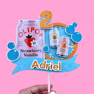 May include: A blue and orange cake topper with the number 2, the name Adriel, and a design featuring cans of Olipop sparkling probiotic drinks. The cans are labeled Strawberry Vanilla, Ginger Lemon, and Orange Squeeze.